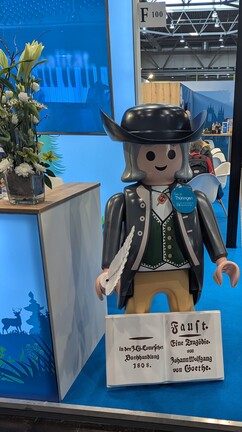 Johann Wolfgang von Goethe als Playmobil-Figur in historischem Gewand mit Schreibfeder und einem Buch, welches einen Auszug aus der ersten Ausgabe von „Faust“ enthält