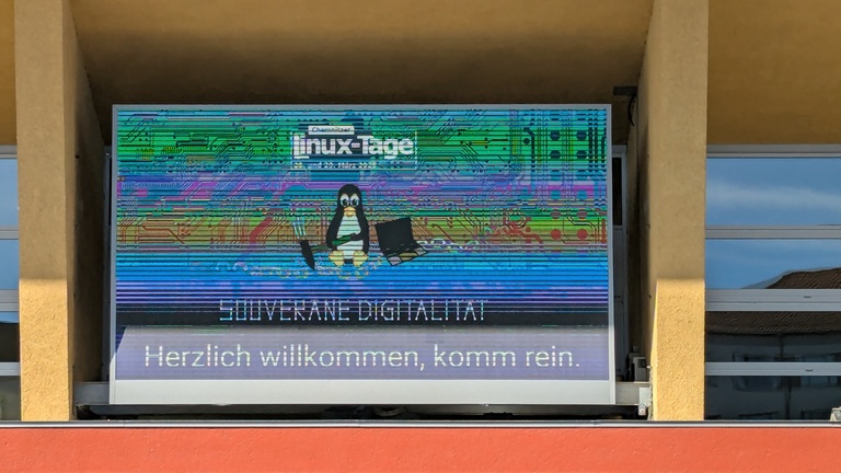 Willkommensbildschirm der Chemnitzer Linux-Tage 2026 über dem Haupteingang des zentralen Hörsaal- und Seminar-Gebäudes der TU Chemnitz, Motto: „Souveräne Digitalität“