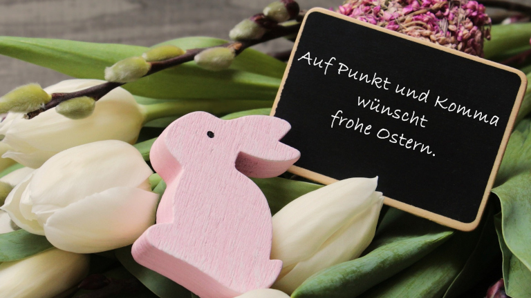 Ein rosa Osterhase aus Holz liegt neben einem Strauß weißer Tulpen, Weidenkätzchen und einer kleinen Tafel, auf der steht: „Auf Punkt und Komma wünscht frohe Ostern.“