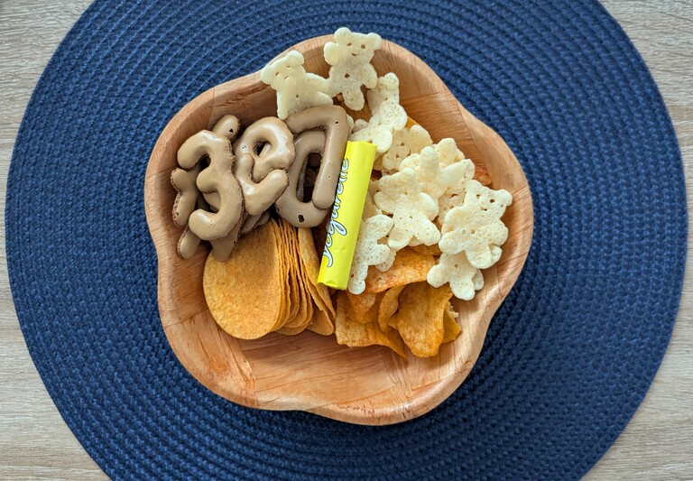 Eine Holzschale mit Chips, Schokolade und Russisch-Brot-Buchstaben auf einem blauen Platzset, bereit für das ESC-Finale.