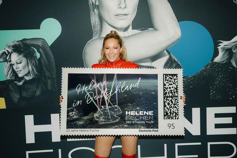 Helene Fischer hält eine überdimensionierte Briefmarke in den Händen, auf der „Helene in Deutschland“ steht.