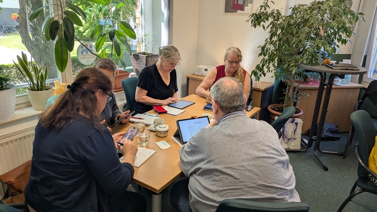 Mehrere Personen sitzen an einem Tisch in einem Raum und arbeiten konzentriert an ihren Tablets. Im Hintergrund stehen Grünpflanzen.