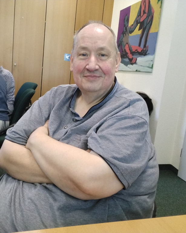 Porträtfoto eines Mannes. Er sitzt auf einem Stuhl an einem Tisch in einem Raum und trägt ein graues Hemd. Die Arme hat er vor sich verschränkt, er lächelt freundlich. Im Hintergrund sind Schränke, ein Bild an der Wand und andere Personen an einem weiteren Tisch zu sehen.
