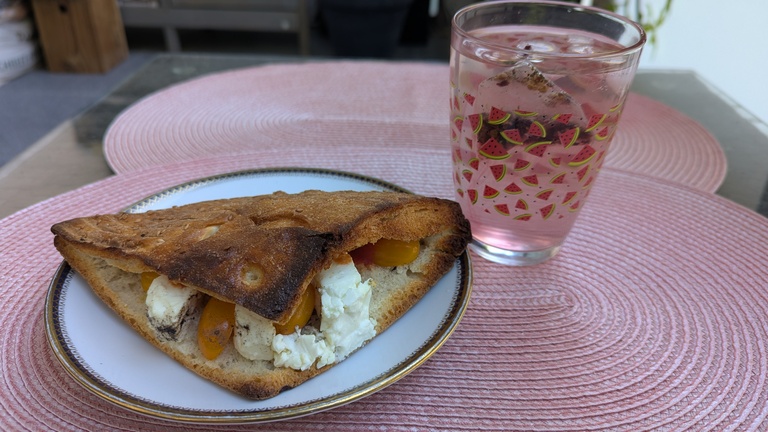 Schafskäse-Fladenbrot liegt auf einem weißen Teller mit schwarz-goldenem Rand. Daneben steht ein Glas mit Wassermelonenmuster, in dem Eiswürfel in Eistee schwimmen. Teller und Glas stehen auf einem rosafarbenen Platzset.
