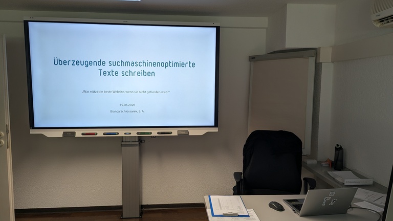 Ein Smartboard und ein Flipchart in einem Schulungsraum. Auf dem Schreibtisch steht ein aufgeklapptes Notebook.