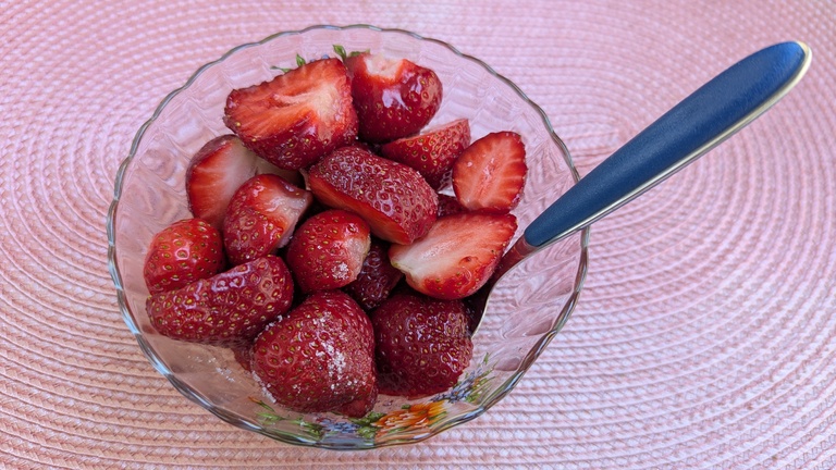 Eine Glasschale voller frischer, halbierter Erdbeeren, die mit feinem Zucker bestreut sind. In der Schale steckt ein Löffel mit dunkelblauem Griff. Das Ganze steht auf einem rosafarbenen Platzset.