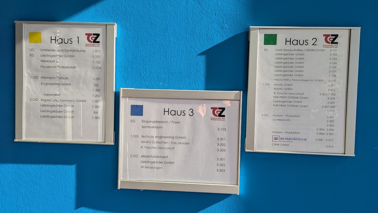 Drei wegweisende Infotafeln an einer hellblauen Wand.
