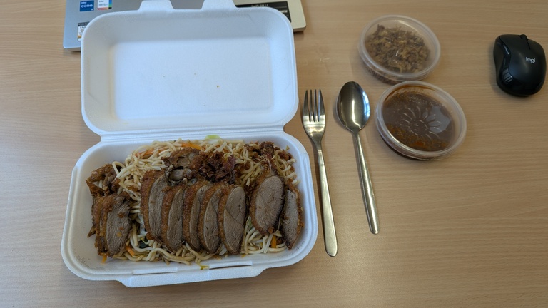 Eine aufgeklappte weiße Box mit gebratenen Nudeln und knuspriger Ente steht auf einem Tisch. Rechts daneben liegen eine Gabel und ein Löffel. Auf dem Tisch befinden sich außerdem je eine Dose mit Röstzwiebeln und scharfer Soße, ein aufgeklapptes Notebook und eine PC-Maus.
