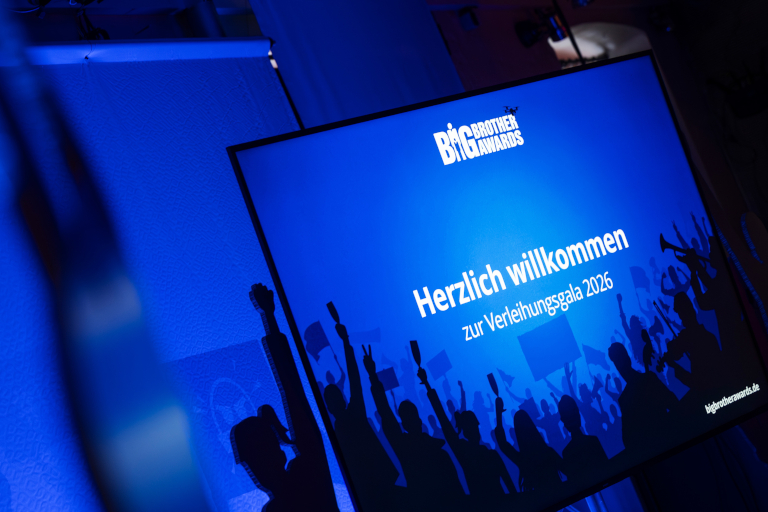 Schräg fotografierter Bildschirm mit blauem Hintergrund und der Aufschrift „Herzlich willkommen zur Verleihungsgala 2026“; darüber das Logo „Big Brother Awards“. Davor sind schwarze Silhouetten einer jubelnden Menschenmenge zu sehen.
